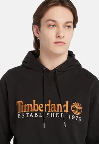 Felpa nera in cotone, con logo "Timberland" ricamato arancione, "FONDATO NEL 1973" e grafica di un albero sul davanti.