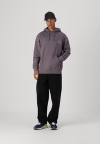 Sudadera gris con capucha y un bolsillo frontal, que presenta un logo en forma de triángulo. Combinada con pantalones negros de pierna ancha y zapatillas oscuras.