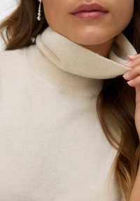 Beige coltrui met een geribbelde textuur, voorzien van een opgevouwen kraag en gemaakt van een zacht, warm materiaal. Lang haar gedeeltelijk zichtbaar.