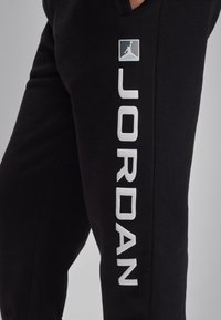 Calças de fato de treino pretas da Jordan com um destaque para o logótipo branco "JORDAN" ao longo da lateral e um pequeno emblema perto da cintura. Textura de tecido macio.