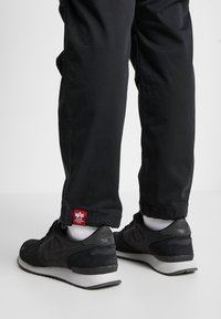 Sorte Nike sneakers har en overdel i ruskind og mesh, polstret krave og en grå gummisål. Bukserne er sorte med et rødt Alpha-label ved kanten.