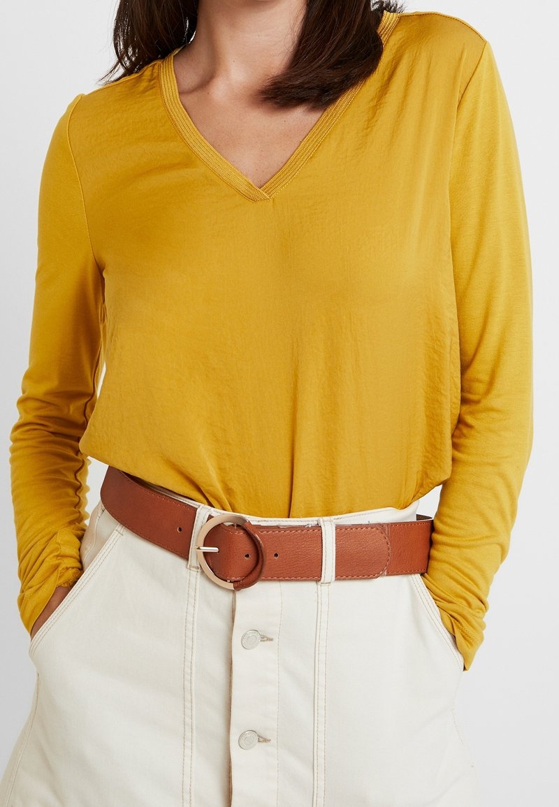 Vrouw in een mosterdgele lange mouwen V-hals top, gestoken in een hooggetailleerde beige rok met knopen aan de voorkant en een bruine riem.