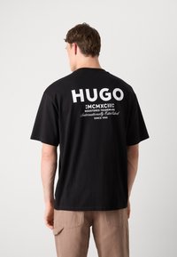 T-shirt in cotone nero con grande testo e logo "HUGO" bianchi stampati sul retro. Vestibilità rilassata e maniche corte, abbinato a pantaloni beige.