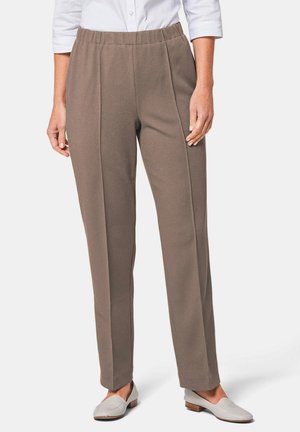 Frau trägt taupefarbene, gerade geschnittene Hose mit vorderen Falten, weiße Loafers und ein weißes Hemd mit Knöpfen und hochgekrempelten Ärmeln.