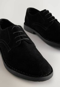 Zapatos de ante negro con cordones, de textura suave, punta redondeada y costuras en tono. Suela de goma con un ligero tacón, diseño minimalista.