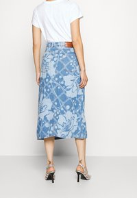 Falda midi de mezclilla con un patrón de flores y lunares en azul claro, cuenta con bolsillos y una etiqueta de cuero en la cintura. Combinada con sandalias de tacón plateado.