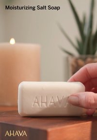 Rektangulär vit tvål med präglad "AHAVA"-logotyp. Slät yta, placerad på en träyta med ett mjukt ljus i bakgrunden.