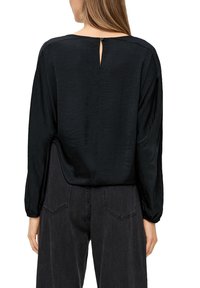 Schwarze Bluse mit langen Ärmeln aus strukturiertem Stoff, Rundhalsausschnitt und einer Schlüssellochausschnitt auf der Rückseite. Kombiniert mit hochgeschnittenen schwarzen Hosen.