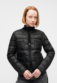 Νεαρό άτομο με κοντά κόκκινα μαλλιά, ντυμένο με μαύρο πουπουλένιο μπουφάν και μαύρο ζιβάγκο, στέκεται μπροστά σε έναν απλό φωτεινό φόντο.