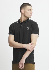 Černé polo tričko s bílými detaily, kr�átkými rukávy a logem na hrudi. Materiál vypadá hladce a přiléhavě, s klasickým designem límce.