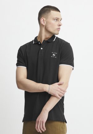 Crna majica polo s bijelim naglascima, kratkim rukavima i logom na prsima. Tkanina izgleda glatko i prilagođeno, s klasičnim dizajnom ovratnika.