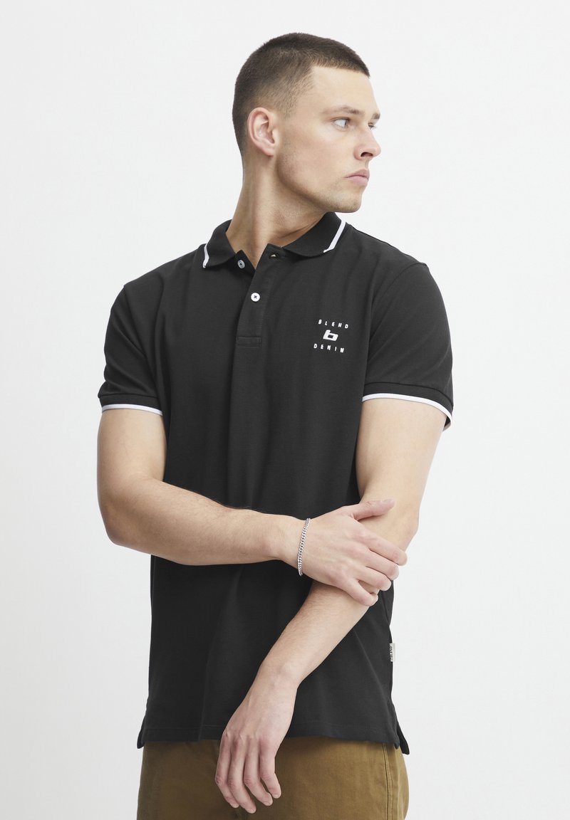 Černé polo tričko s bílými detaily, krátkými rukávy a logem na hrudi. Materiál vypadá hladce a přiléhavě, s klasickým designem límce.