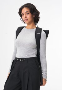 Schwarzer Rucksack mit verstellbaren Trägern, getragen über einem hellgrauen Langarmshirt. Locker sitzende schwarze Hose mit Falten und einer Schnallendetail.