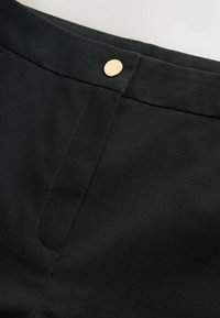 Détail de la taille d'un pantalon noir avec un bouton doré et une fermeture éclair dissimulée sur la braguette.