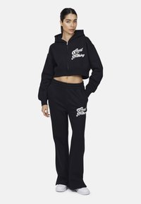 Zwarte cropped hoodie met een rits aan de voorkant en de tekst "Good for Nothing". Gecombineerd met losse zwarte broek met hetzelfde logo. Soepele textuur.
