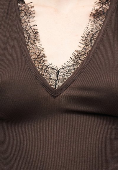 Καφέ ριμπ πλεκτό τοπ με V-λαιμόκοψη, φορεμένο πάνω από μαύρη διακοσμημένη δαντέλα με λουλούδια, που αποκαλύπτει το άνω μέρος του στήθους και την περιοχή της κλείδας.
