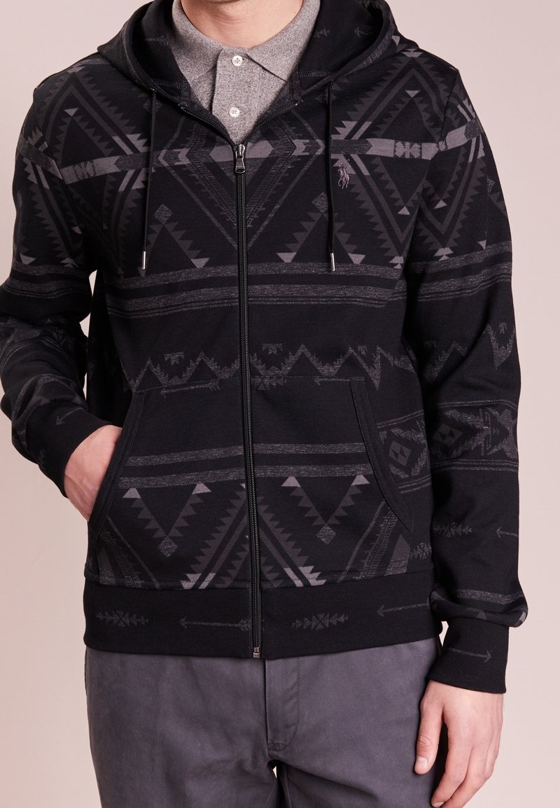 Svart zip-up hoodie med ett intrikat mörkgrått geometriskt mönster, som har en huva, sidofickor och ribbade ärmslut och nederkant.