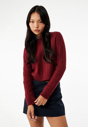Ung kvinde med langt sort hår iført en rød kabelstrikket sweater og sort nederdel foran en ensfarvet hvid baggrund.