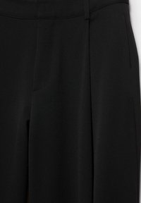 Gros plan sur un pantalon noir sur mesure avec un pli frontal et une ceinture avec passants.