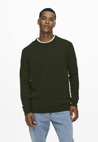 Suéter verde oscuro con cuello redondo y mangas largas, con puños y dobladillo acanalados, usado sobre unos jeans de color azul claro.