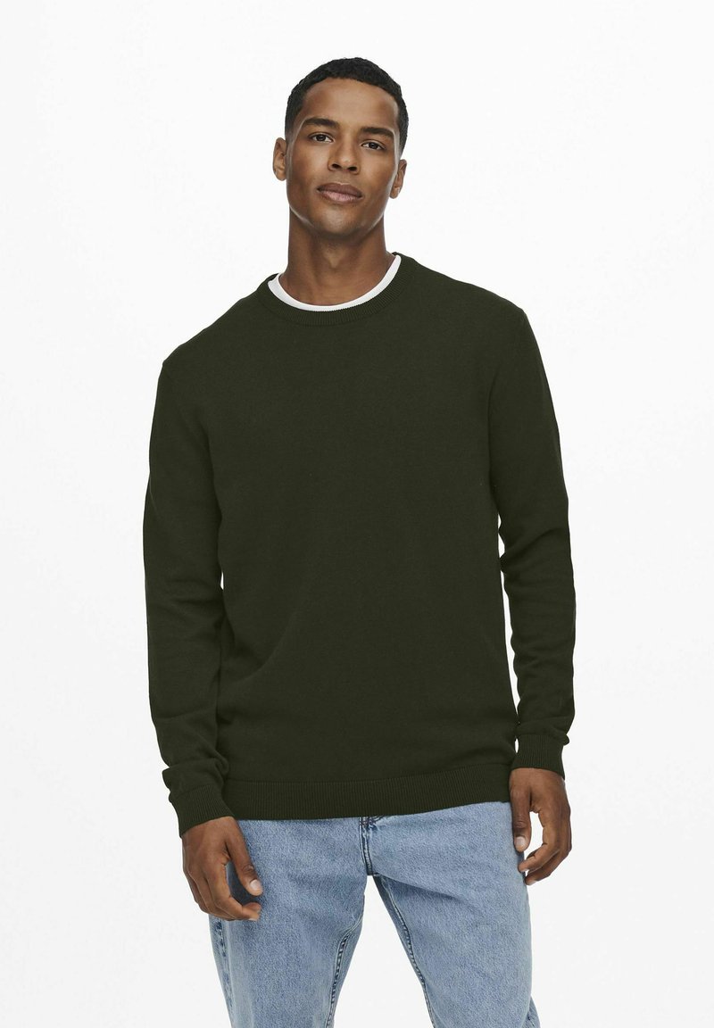 Suéter verde oscuro con cuello redondo y mangas largas, con puños y dobladillo acanalados, usado sobre unos jeans de color azul claro.
