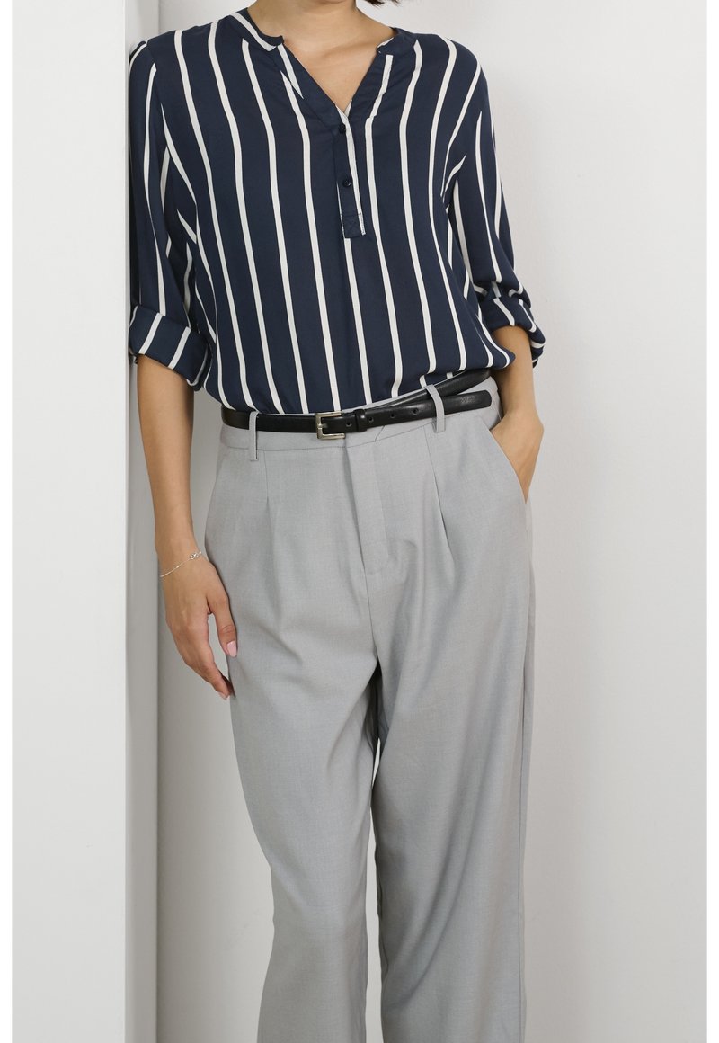 Blouse rayée bleu marine et blanche avec un col en V, manches retroussées, assortie à un pantalon taille haute gris clair et une ceinture noire.