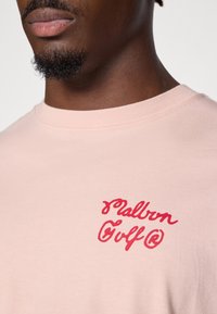 Lyserød bomulds t-shirt med rund halsudskæring og korte ærmer, der prydes af rød broderet "Malbon Golf" tekst på venstre brystområde.