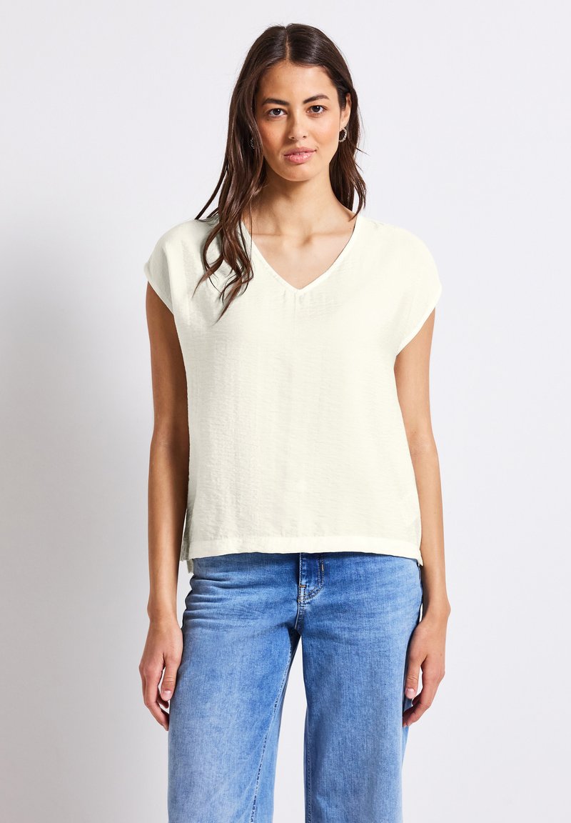V-Ausschnitt, kurzärmliges Oberteil in hellem Creme, aus leichtem strukturiertem Stoff, lässige Passform, kombiniert mit ausgewaschenen blauen Jeans.