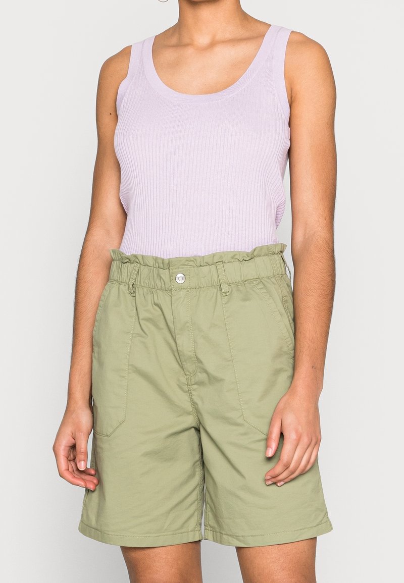 Femme portant un débardeur côtelé violet clair et un short taille haute vert olive à effet papier, debout devant un fond uni.
