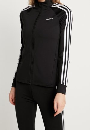 Vrouw draagt een zwart Adidas trainingsjack met witte strepen op de mouwen en bijpassende zwarte legging met witte strepen.