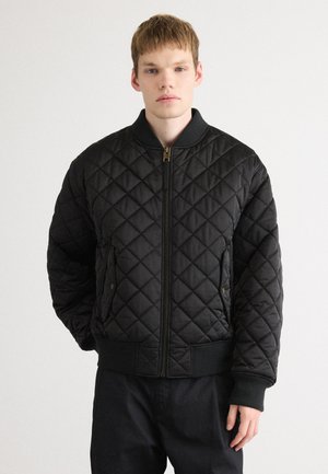 Young man wearing a black quilted bomber jacket with ribbed collar, cuffs, and hem, standing against a plain light background.