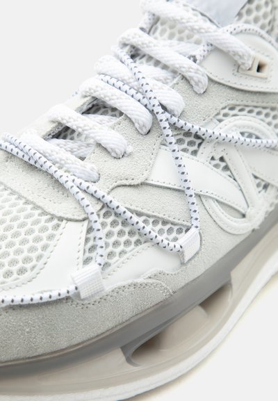 Κοντινή λήψη λευκού και γκρι αθλητικού παπουτσιού με πάνελ από δίχτυ και σουέντ, λευκά κορδόνια με σχέδιο και ορατό τμήμα της σόλας με αεροθαλάμο.