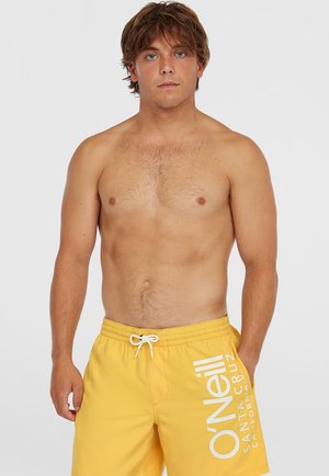 Yellow swim trunks with white logo, elastic waistband, and drawstring. Worn by a person with short hair and a neutral expression.