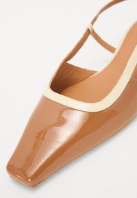 Beige kiiltonkainen slingback-kengen, jossa on neliön muotoinen kärki, ja jonka ylhäällä on kontrastivärinen cream-värinen reuna ja ohut nilkkahihna.