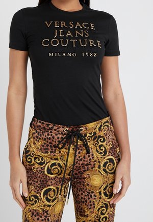 Czarny T-shirt ze złotym napisem "Versace Jeans Couture Milano 1988", zestawiony z legginsami w panterkę zdobionymi ozdobnymi złotymi barokowymi wzorami.