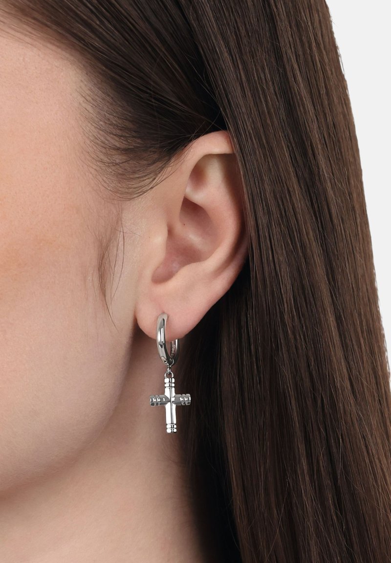 Silberner Creolenohrring mit einem hängenden Kreuzdesign, das eingravierte Details und eine polierte Oberfläche aufweist. Trägt ein Modell mit glattem Haar am Ohr.