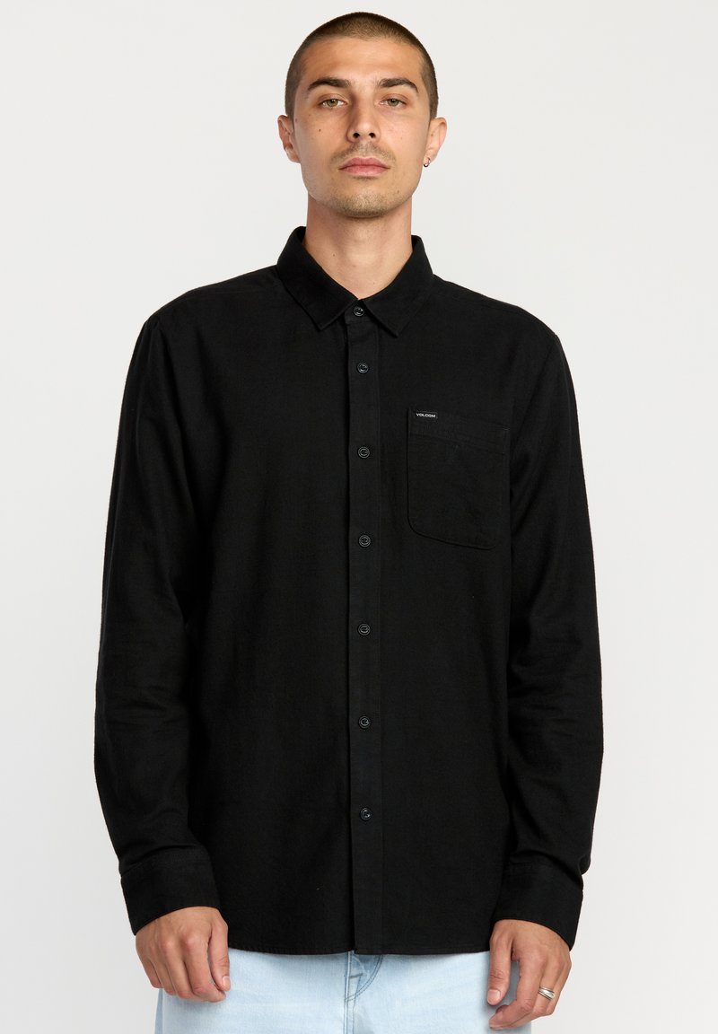 Chemise noire à manches longues en coton, avec un col boutonné, une poche avant et une étiquette de marque sur la poitrine. Texture lisse et design ajusté.