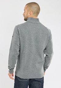 Pull en maille gris avec un col montant et des poignets côtelés, présentant une texture subtile. Porté par-dessus un jean bleu marine, vu de dos.