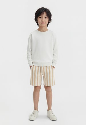 Garçon debout les mains dans les poches, portant un sweat-shirt blanc, un short à rayures verticales beige et blanches, et des baskets blanches sur fond uni.