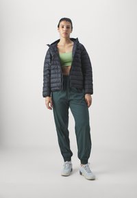 Padded schwarze Jacke, grünes Cropped Top und dunkelgrüne Jogginghosen mit elastischen Bündchen. Graue Schuhe mit strukturierten Sohlen. Einfaches Design, keine sichtbaren Muster.