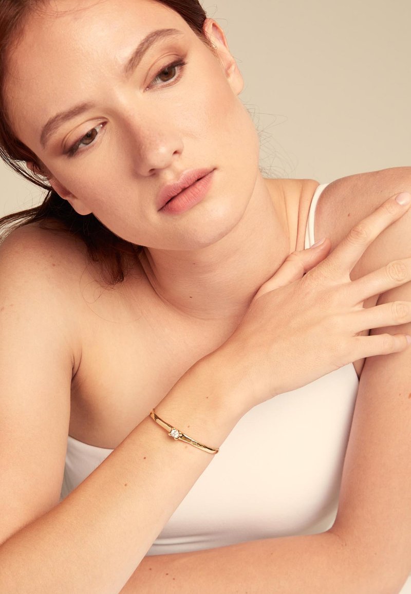 Pulsera de oro con un diseño simple, que presenta un pequeño acento de diamante. El brazo del modelo es parcialmente visible, mostrando una piel suave y una iluminación suave.