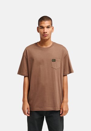 Uomo che indossa una maglietta marrone larga con una piccola toppa logo nera e gialla sulla tasca del petto, abbinata a pantaloni scuri.