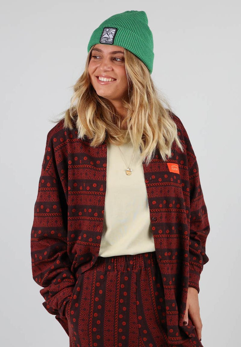 Veste rouge à motifs sur un top crème, associée à un pantalon rouge à motifs. Un bonnet vert repose sur de longs cheveux ondulés.