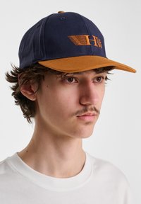 Navy baseball cap with a brown visor, featuring an embroidered logo in orange. The fabric appears smooth with visible stitching details.