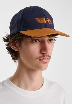 Navy baseball cap with a brown visor, featuring an embroidered logo in orange. The fabric appears smooth with visible stitching details.