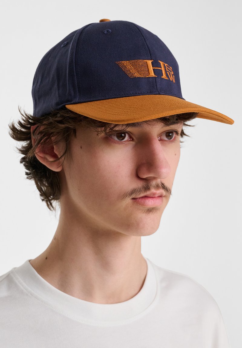Navy baseball cap with a brown visor, featuring an embroidered logo in orange. The fabric appears smooth with visible stitching details.