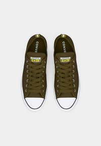 Un paio di sneakers Converse All Star verde oliva con punte e lacci bianchi, mostrati dall'alto su uno sfondo bianco.