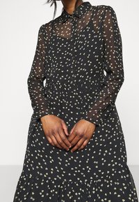 Svart klänning med genomskinliga långärmar, knäppning fram och lager-på-lager kjol. Vita och gula lövmönsteraccenter genom hela plagget.