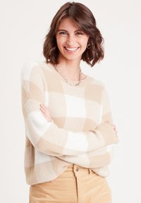 Beige-weiß kariertes Sweatshirt mit weichem Material, lockerem Schnitt und langen Ärmeln, kombiniert mit hellen Cordhosen. Silberne Kettenhalskette.