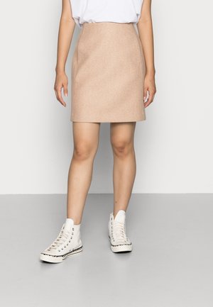 Femme portant une jupe beige longueur genoux, une chemise blanche rentrée et des baskets blanches montantes, debout sur un sol gris contre un mur blanc.